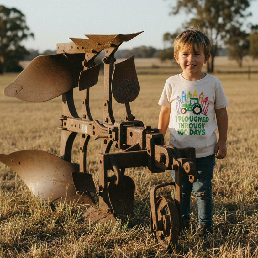 A young boy stands in a dry grass field next to a large rusted agricultural plow, wearing a white graphic t-shirt with a colorful tractor and the text 'I PLOUGHED THROUGH 100 DAYS'.
