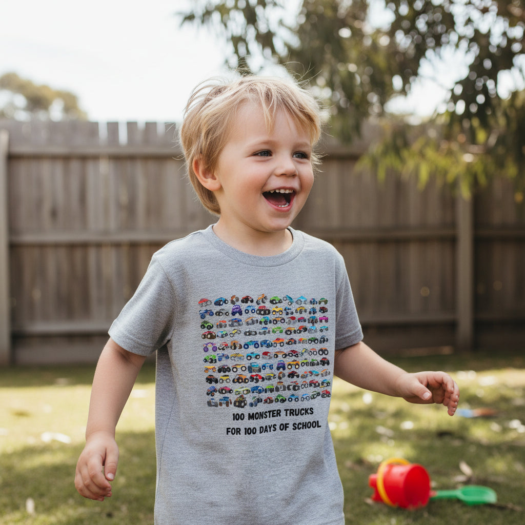 A young blonde boy wearing a light gray graphic t-shirt with a colorful monster truck design and the text '100 MONSTER TRUCKS FOR 100 DAYS OF SCHOOL', standing outdoors in a backyard with a wooden fence and grass.
