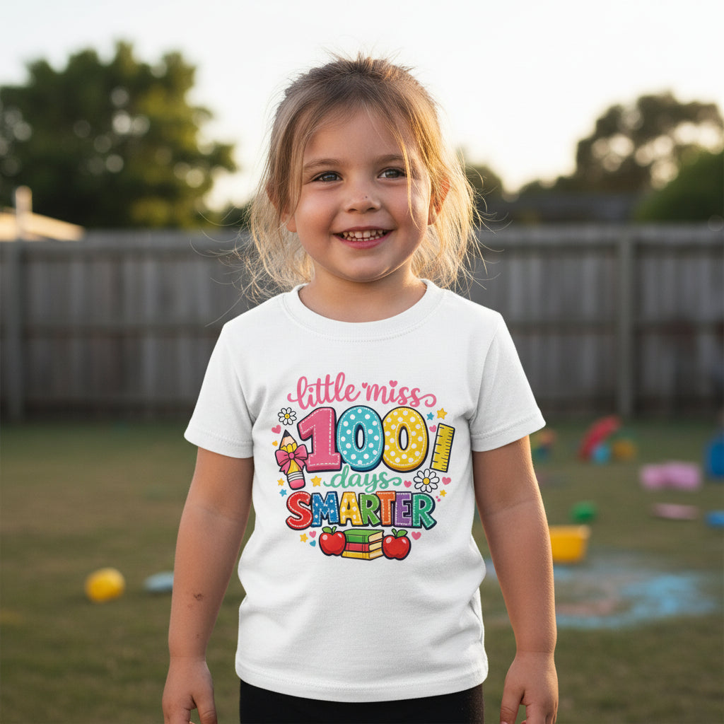 A young girl smiling in a backyard wearing a white graphic t-shirt that reads 'little miss 100 days SMARTER' with colourful school-themed illustrations.