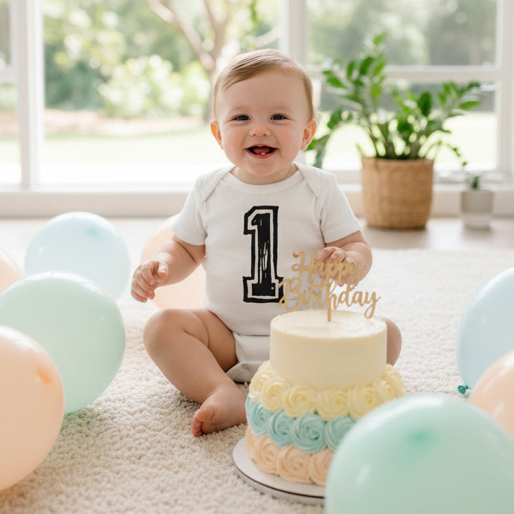 Baby wearing a white bodysuit with a black number 1 design, sitting beside a first birthday cake.