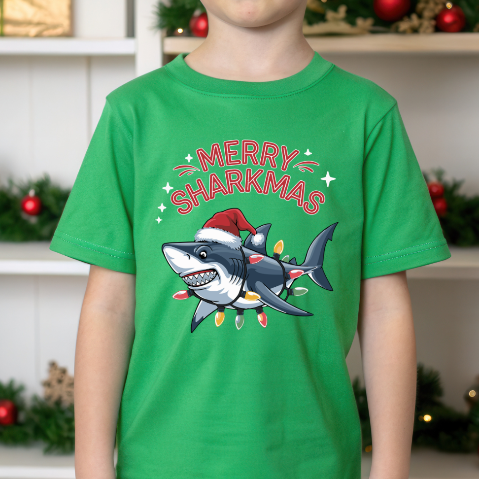 Child wearing a green t-shirt with a shark wearing a Santa hat and 'Merry Sharkmas' text, Christmas decorations in the background.