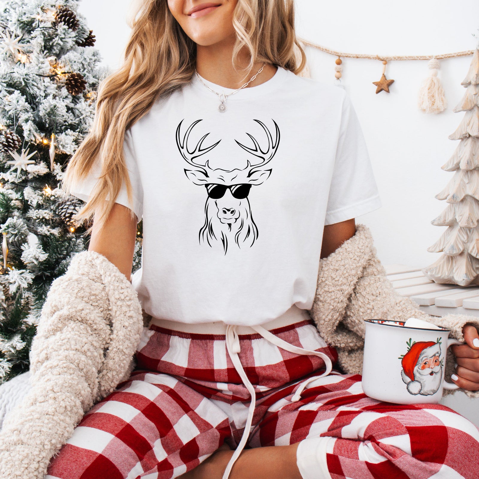 A person wearing a white Christmas-themed t-shirt with a reindeer and sunglasses design, sitting in front of a blurred Christmas tree and decorations.