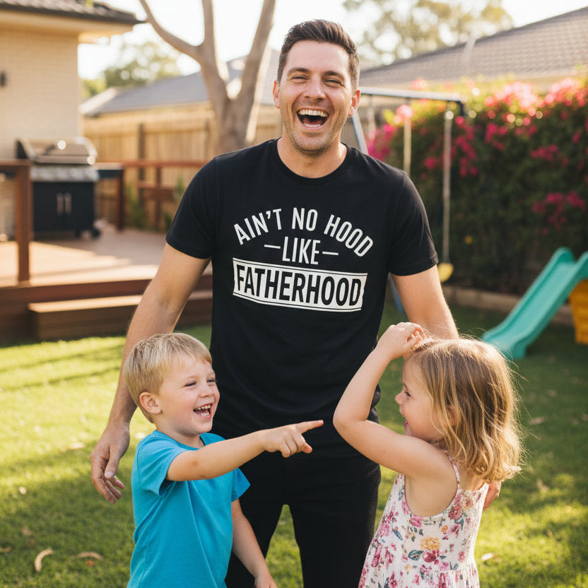Dad wearing black Ain't No Hood Like Fatherhood t-shirt laughing with two children in backyard