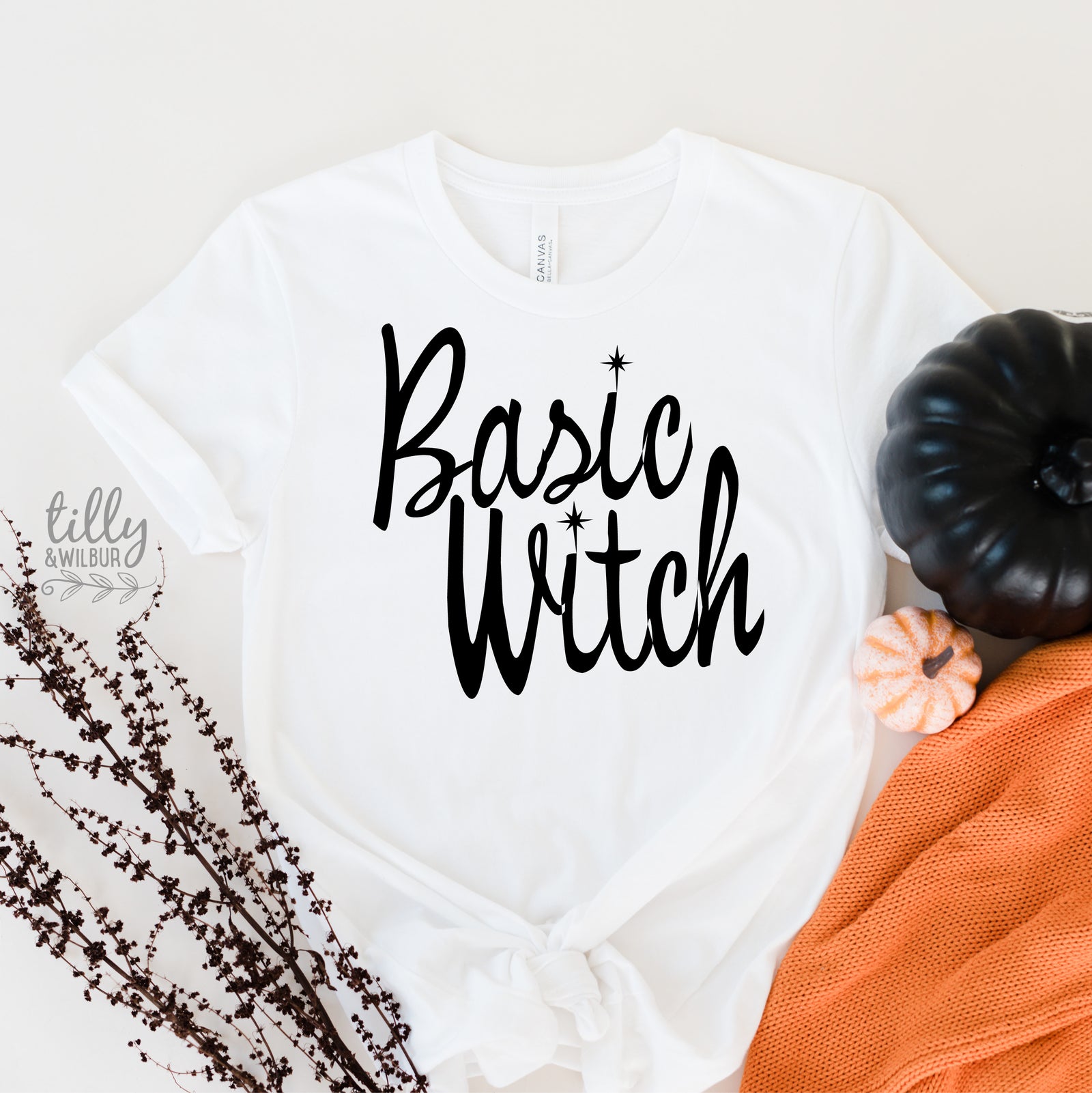A white graphic T-shirt laid flat with the words 'Basic Witch' printed in large black script across the front. The shirt is styled with a small orange pumpkin, a black pumpkin, dried branches, and an orange knit fabric on a light neutral background.