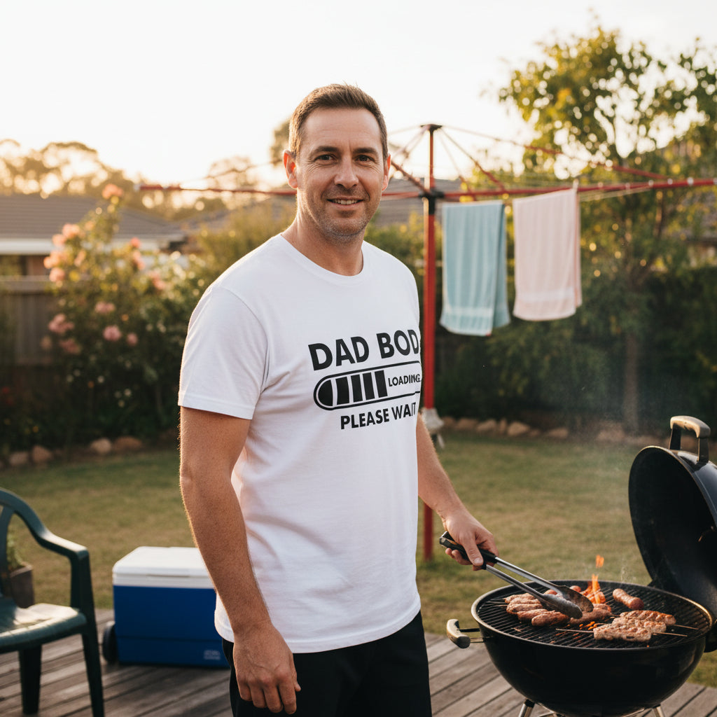 A man wearing a white graphic t-shirt with 'DAD BOD LOADING PLEASE WAIT' and a battery icon, standing next to a charcoal grill in a backyard.