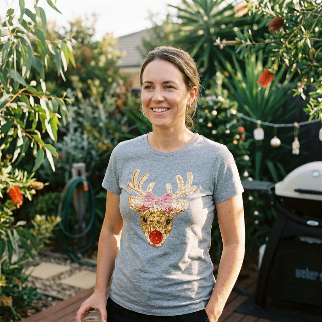 A heather gray short-sleeve t-shirt featuring a glittery gold reindeer graphic with a pink bow and a red nose on the front, displayed against a plain white background.
