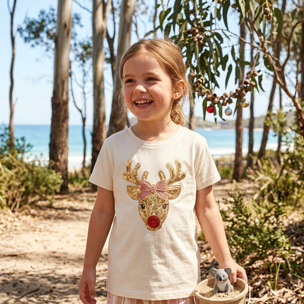 An off-white short-sleeve t-shirt featuring a festive Christmas reindeer graphic on the front. The reindeer has gold glittery antlers and ears, a pink glittery bow, and a red nose, set against a clean white background.