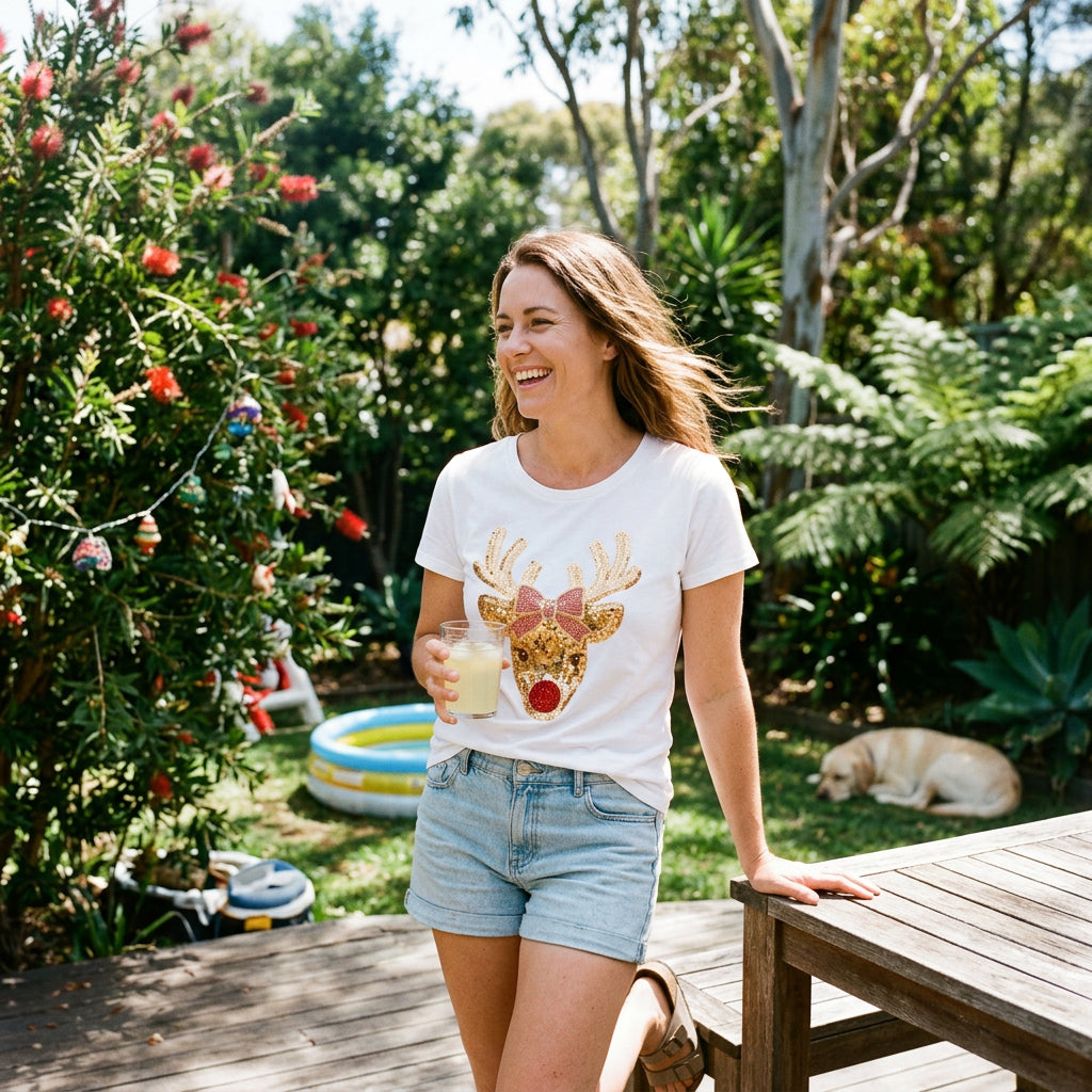 A white short-sleeve crewneck t-shirt featuring a glittery gold reindeer graphic with a pink bow and a red nose on the front, displayed against a solid white background.