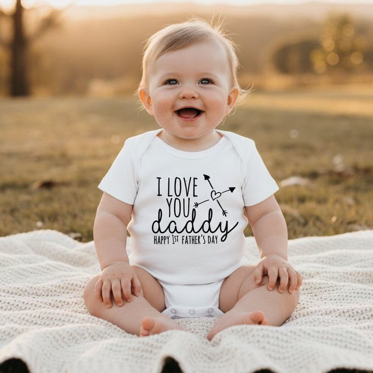A white short-sleeve baby bodysuitprinted with the phrase 'I LOVE YOU daddy HAPPY 1st FATHER'S DAY' in black lettering with a small heart and arrow graphic.