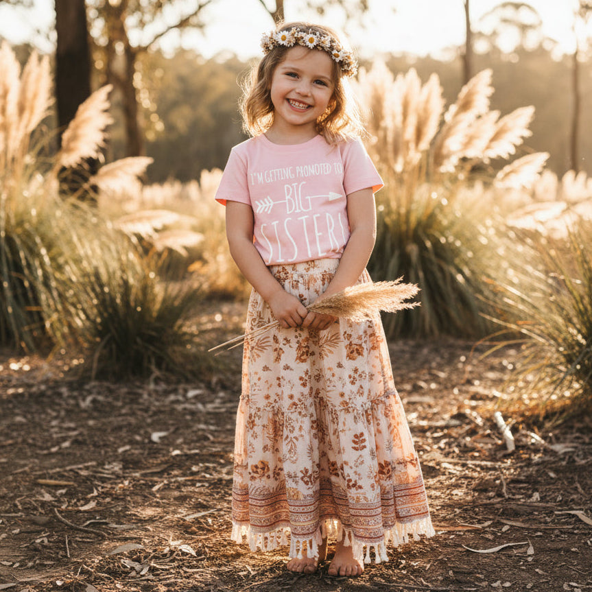 Little girl in a pink "I'm Getting Promoted To Big Sister" tee standing in a sunlit field wearing a daisy crown.