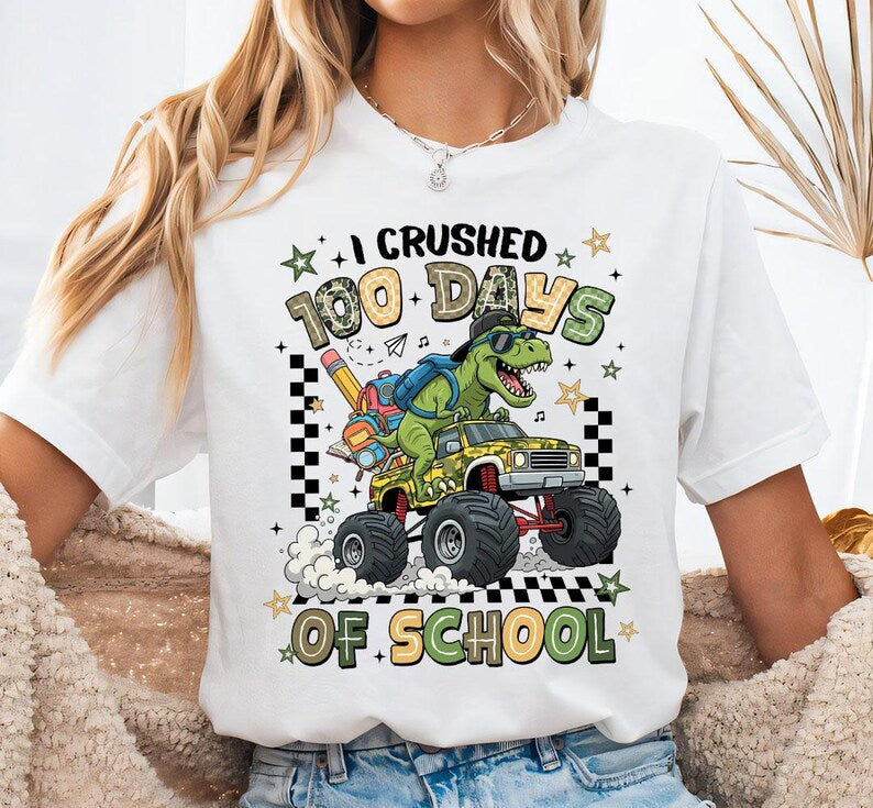 A woman wearing a white graphic t-shirt with a cartoon dinosaur monster truck design and the text 'I CRUSHED 100 DAYS OF SCHOOL'.