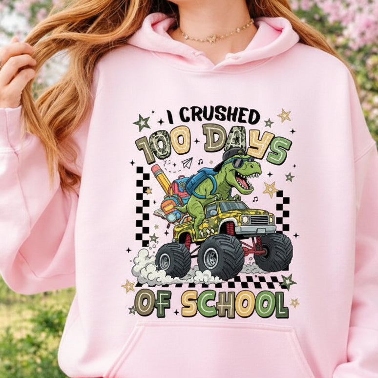 A person wearing a light pink sweatshirt with a colorful graphic that reads 'I CRUSHED 100 DAYS OF SCHOOL' featuring a green dinosaur driving a yellow monster truck. The background is a blurred outdoor garden setting.