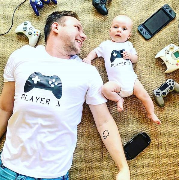 Dad and baby in matching white tops with controller designs reading Player 1 and Player 2.