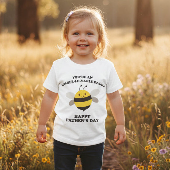 Toddler in a field wearing a white t-shirt with a cartoon bee reading You're An Un-Bee-Lievable Daddy, Happy Father's Day.