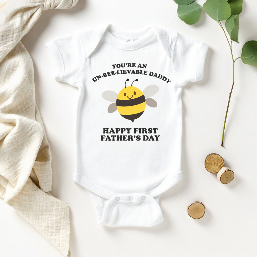 White baby bodysuit with a cartoon bee and text reading You're An Un-Bee-Lievable Daddy, Happy First Father's Day.
