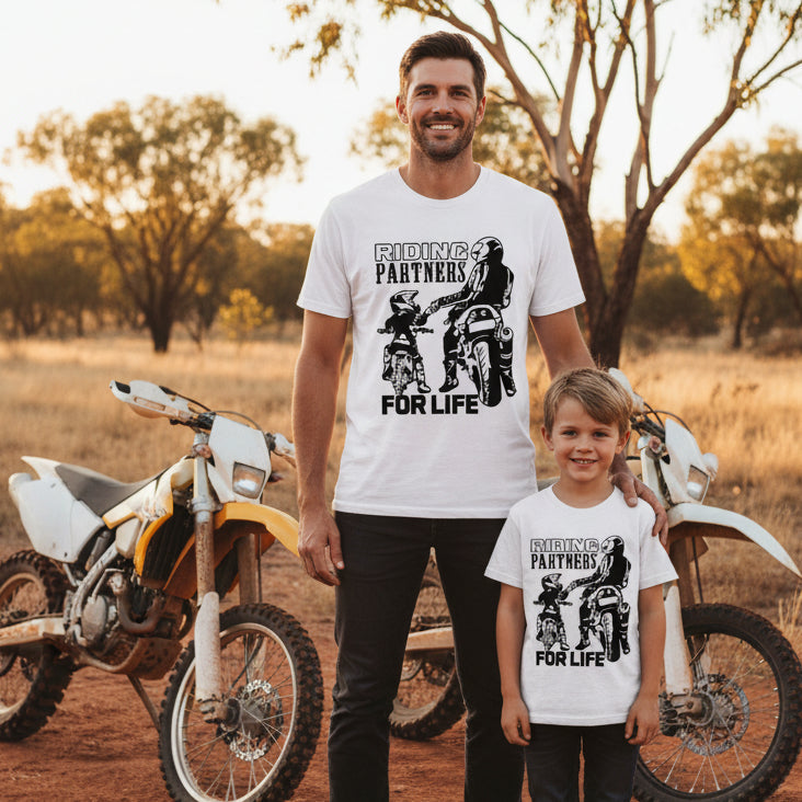 Two white t-shirts laid flat showing matching Riding Partners For Life design with dad and son on motorbikes fist bumping in bold black silhouette