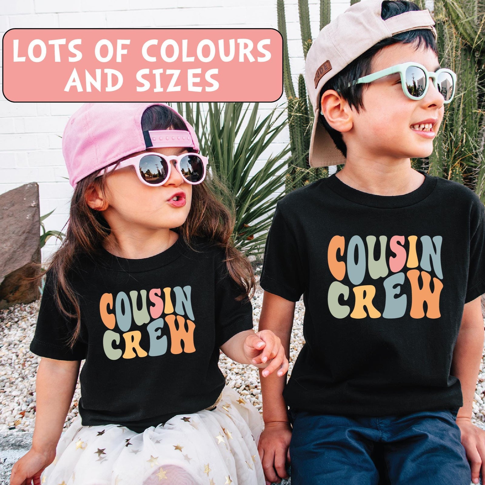 Two children sitting in front of a cactus, wearing black T-shirts with the text ' Cousin Crew ' in colorful letters.