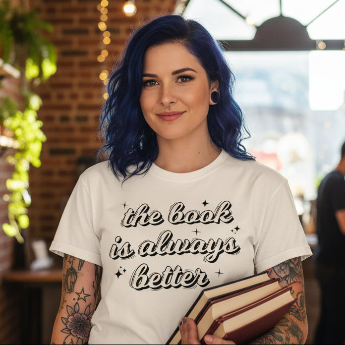 Woman holding books wearing a cream The Book Is Always Better t-shirt with bold retro script slogan and sparkle accents