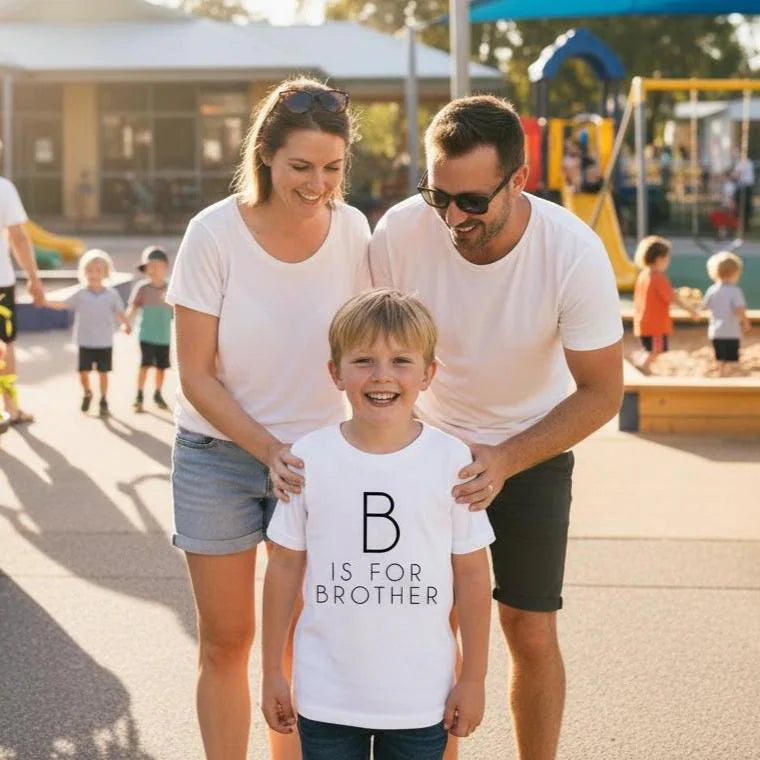 Young boy smiling outdoors wearing a white B Is For Brother t-shirt with minimal monogram design, standing with his parents