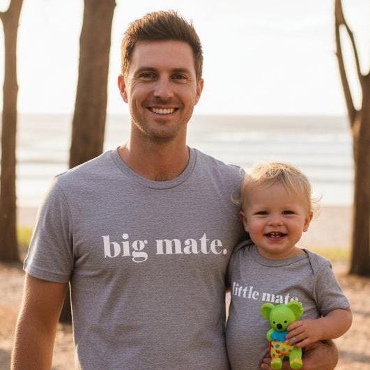 Tilly&Wilbur grey matching set — an adult tee printed with "big mate." and a baby bodysuit printed with "little mate." in minimalist white lowercase text, worn by a dad carrying his baby near the beach.