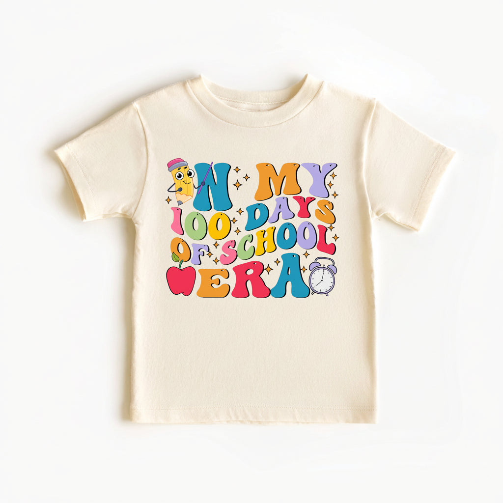 A cream-colored short-sleeve baby t-shirt featuring a colorful retro-style graphic that reads 'IN MY 100 DAYS OF SCHOOL WERA' with cartoon graphics. A pink circular badge in the bottom right corner reads 'LOTS OF SIZES lilly brand & COLOURS'.