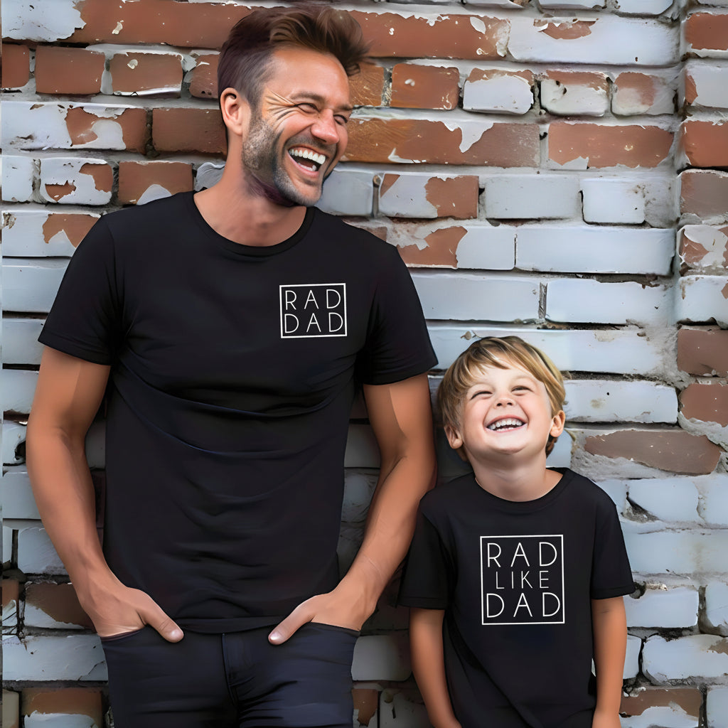 A man and a child wearing matching black t-shirts with a white 'RAD LIKE DAD' graphic on the chest, standing against a brick wall. A white circular badge with the text 'MORE COLOURS' is overlaid on the top right.