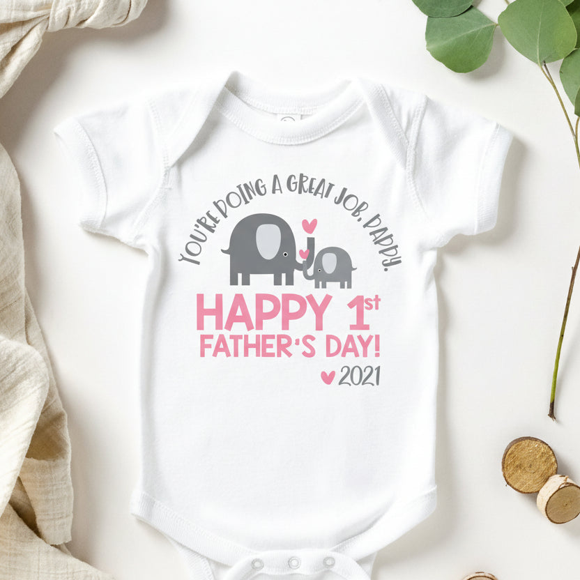 A white short-sleeve baby bodysuit laid flat on a light background, printed with a gray elephant and baby elephant graphic and pink text reading 'You're doing a great job baby Happy 1st Father's Day! 2026.' Decorative beige fabric, green leaves, and small wooden beads surround the bodysuit.