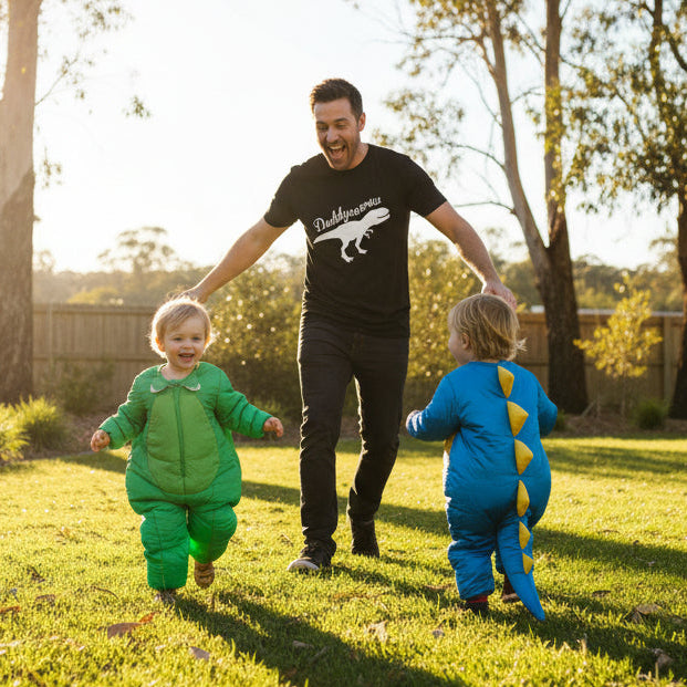A man wearing a black t-shirt with a white dinosaur graphic walks hand-in-hand with two children in dinosaur onesies on a sunny grassy backyard. The child on the left wears a green dinosaur onesie, and the child on the right wears a blue dinosaur onesie with yellow back spikes.