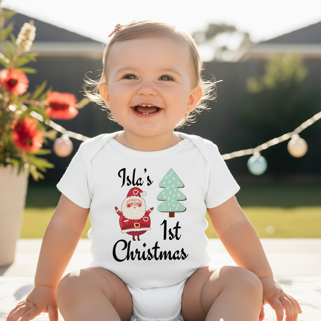 Smiling baby wearing a white personalised “First Christmas” bodysuit with name Isla, Santa illustration and Christmas tree, outdoor setting
