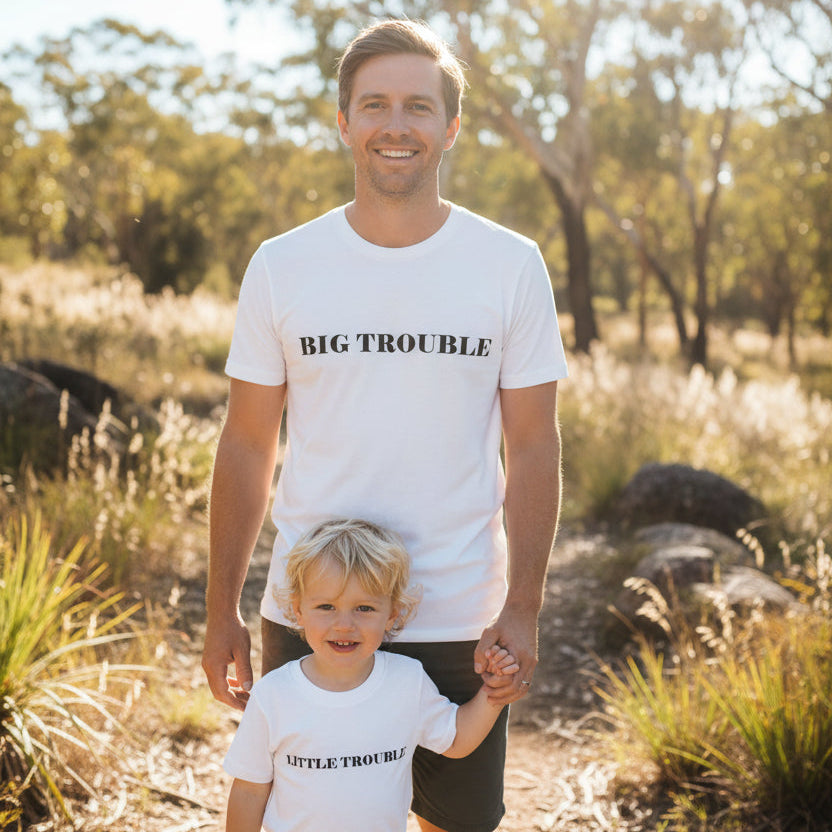 Dad and young son standing in bushland, both wearing matching white t-shirts that read Big Trouble and Little Trouble.