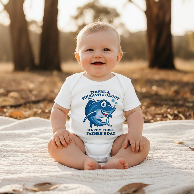White baby bodysuit with a cartoon shark and text reading You're A Fin-Tastic Daddy, Happy First Father's Day.
