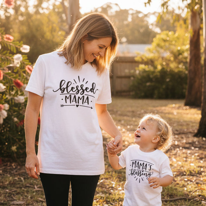 Matching mum and baby outfits with “Blessed Mama” t-shirt and “Mama’s Blessing” baby bodysuit, black text on white garments, flat lay display