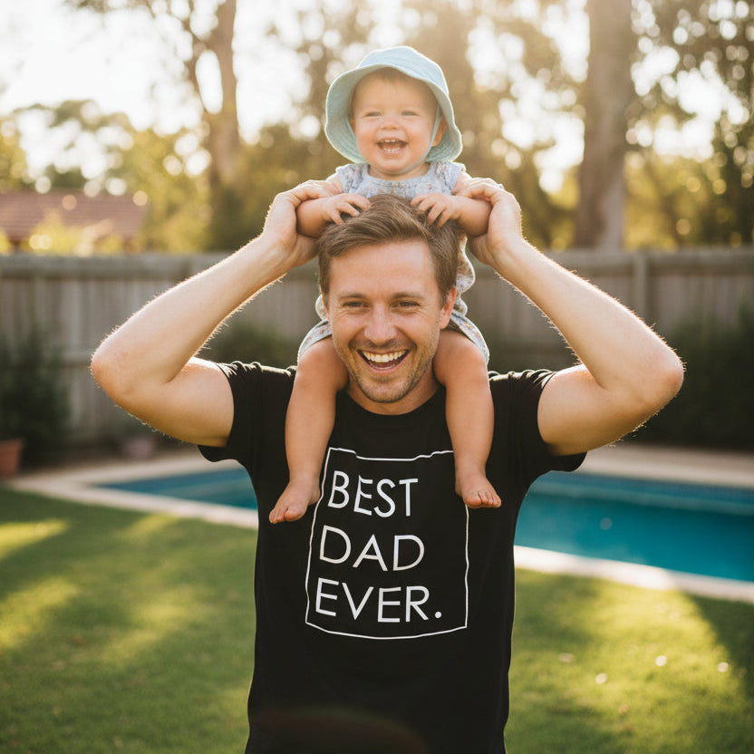 Dad wearing a black Best Dad Ever t-shirt with a smiling baby on his shoulders in a backyard