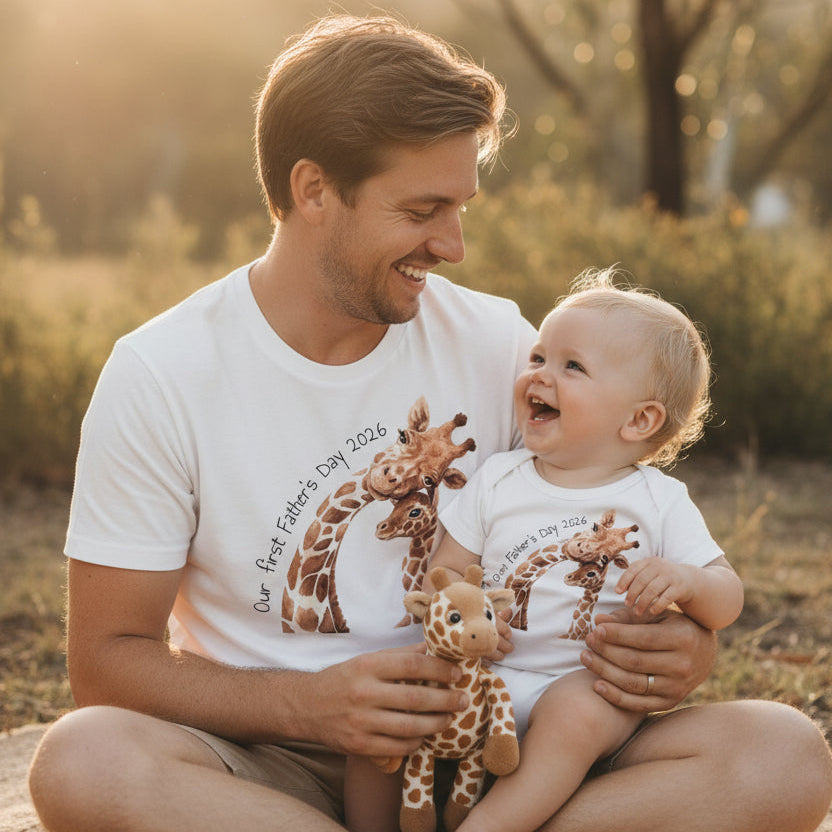 Dad and baby in matching white tops with a giraffe Our First Father's Day design, holding a giraffe toy.
