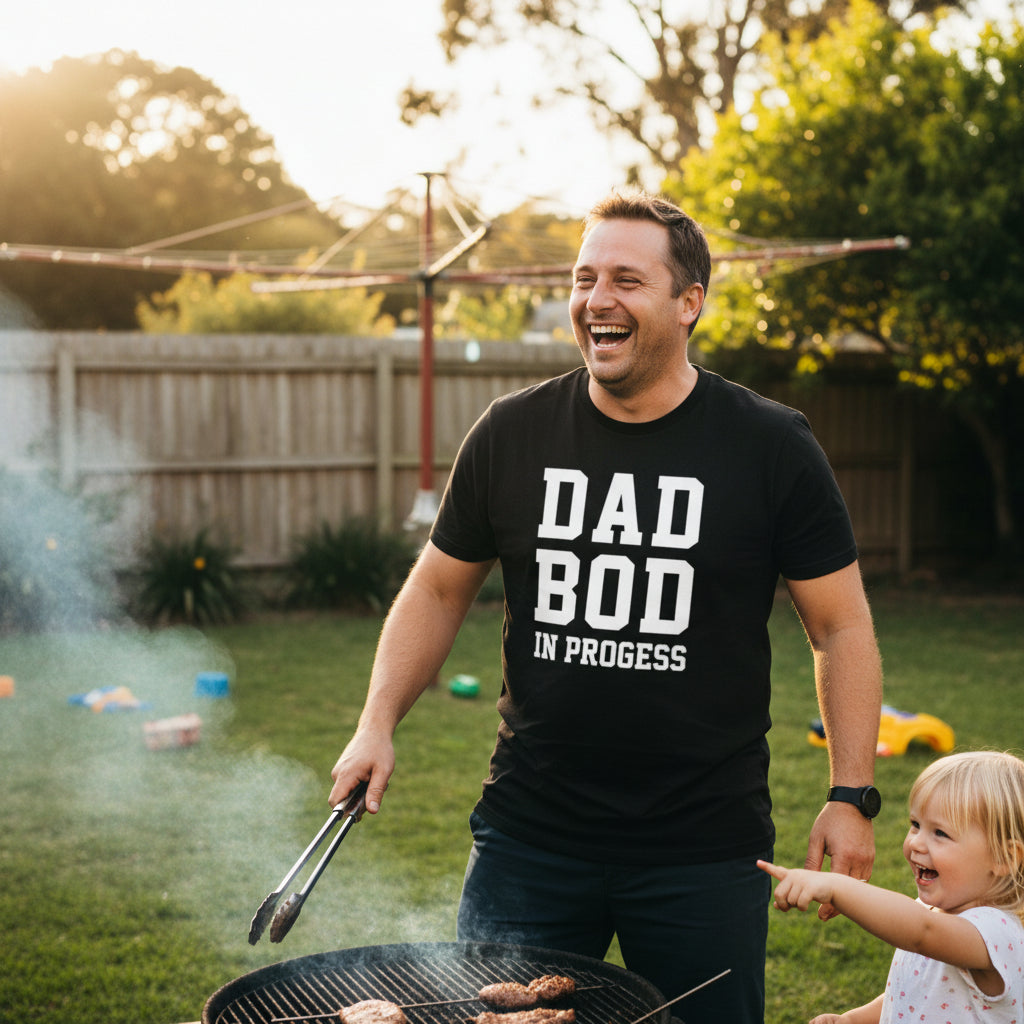 Black t-shirt reading Dad Bod In Progress in bold white varsity lettering.