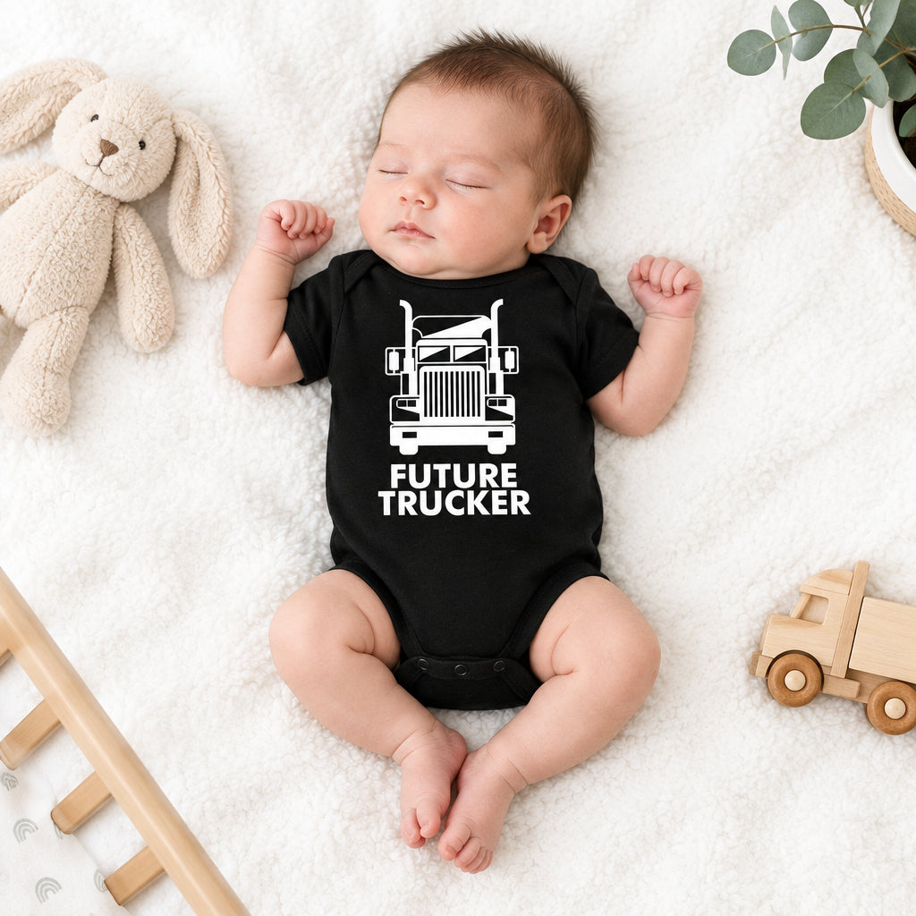 A sleeping newborn baby wearing a black short-sleeve bodysuit with a white truck graphic and the words 'FUTURE TRUCKER,' lying on a white textured blanket with plush toys and a wooden toy truck around.