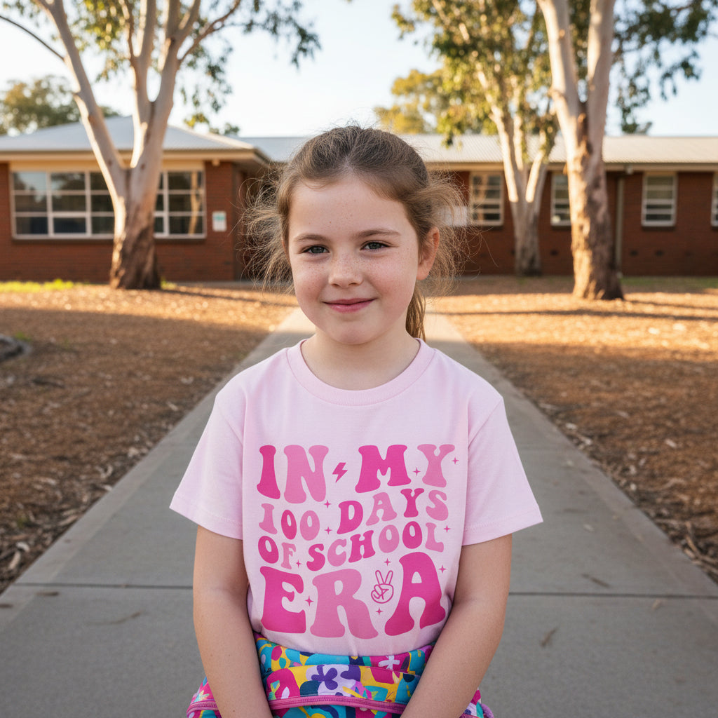A young girl stands on a concrete path in front of a brick school building, wearing a light pink graphic t-shirt that reads 'IN MY 100 DAYS OF SCHOOL ERA' in dark pink and purple lettering, paired with a colorful patterned skirt.