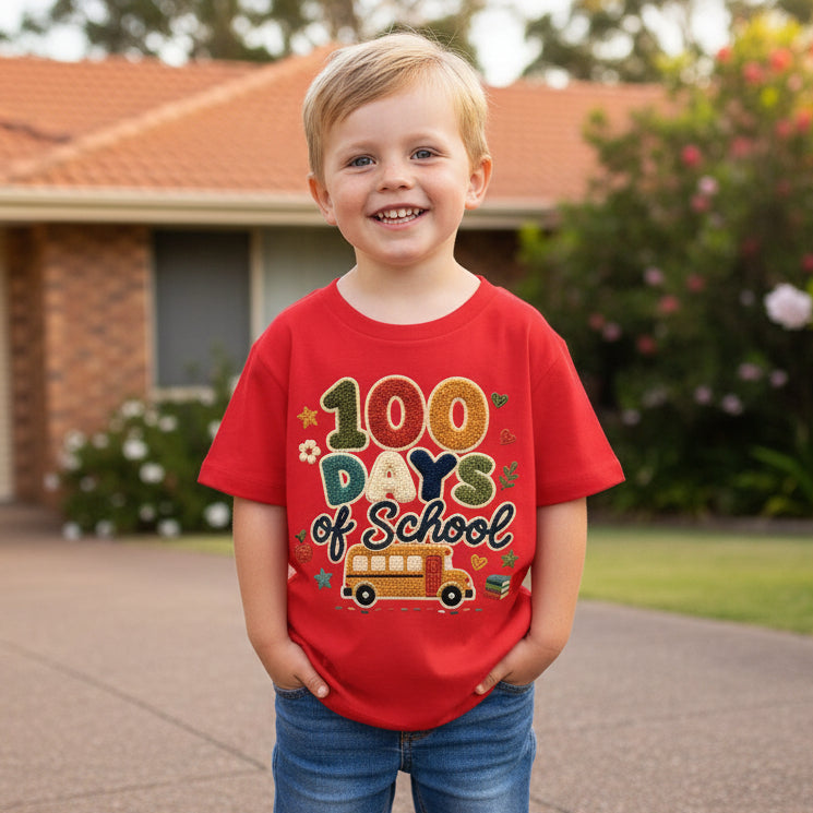 A young boy smiling outdoors in front of a house, wearing a bright red graphic T-shirt that reads '100 Days of School' with a school bus illustration and blue jeans.