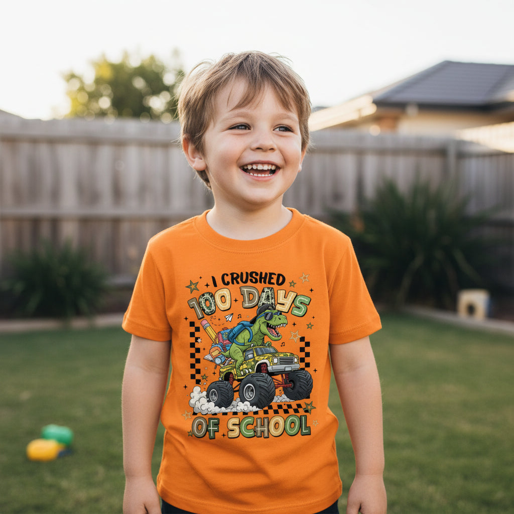 A young boy smiling outdoors in a backyard, wearing a bright orange graphic t-shirt that reads 'I CRUSHED 100 DAYS OF SCHOOL' with a cartoon dinosaur and monster truck design.