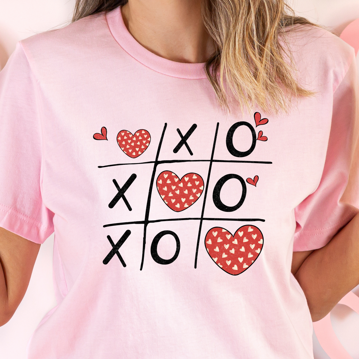 A person wearing a pink t-shirt with a graphic of a &#39;Naughts and Crosses&#39; game board, filled with red hearts and the text &#39;LOLs OF COLOURS &amp; SIZES&#39;.