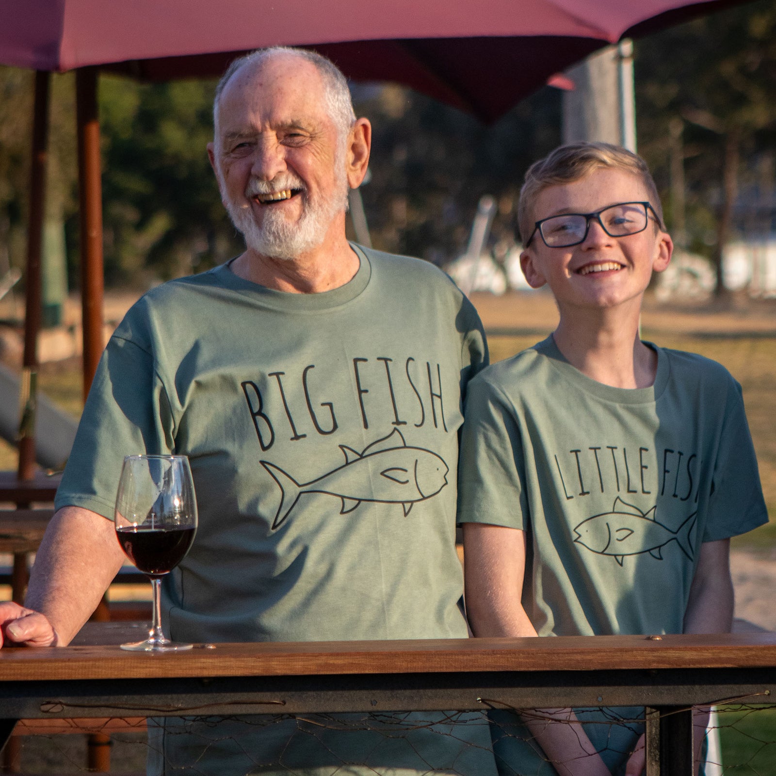 An older man and a young boy smiling, wearing matching green t-shirts with the text 'BIG FISH' and 'LITTLE FISH' respectively, each printed with a graphic of a fish.