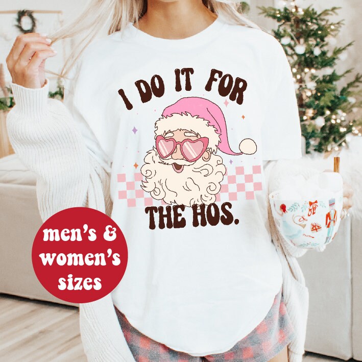 A person wearing a white long-sleeved sweatshirt with a colorful graphic of Santa Claus and the text 'I DO IT FOR THE HO'S' on the front, holding a coffee cup, with a Christmas tree and gifts in the background.