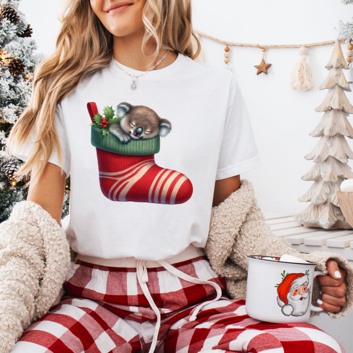 A person wearing a white t-shirt with a colorful christmas themed koala bear design, sitting in a cozy room with a christmas tree and gifts in the background.