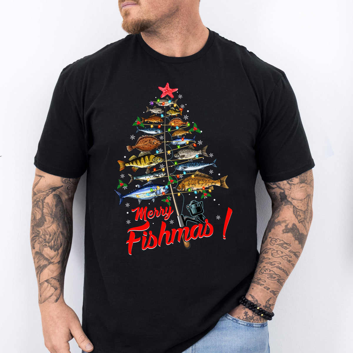 A man wearing a black T-shirt with a colorful Christmas tree design and the text &#39;Merry Fishmas!&#39; on it, standing in a casual outfit with a tattoo visible on his arm.