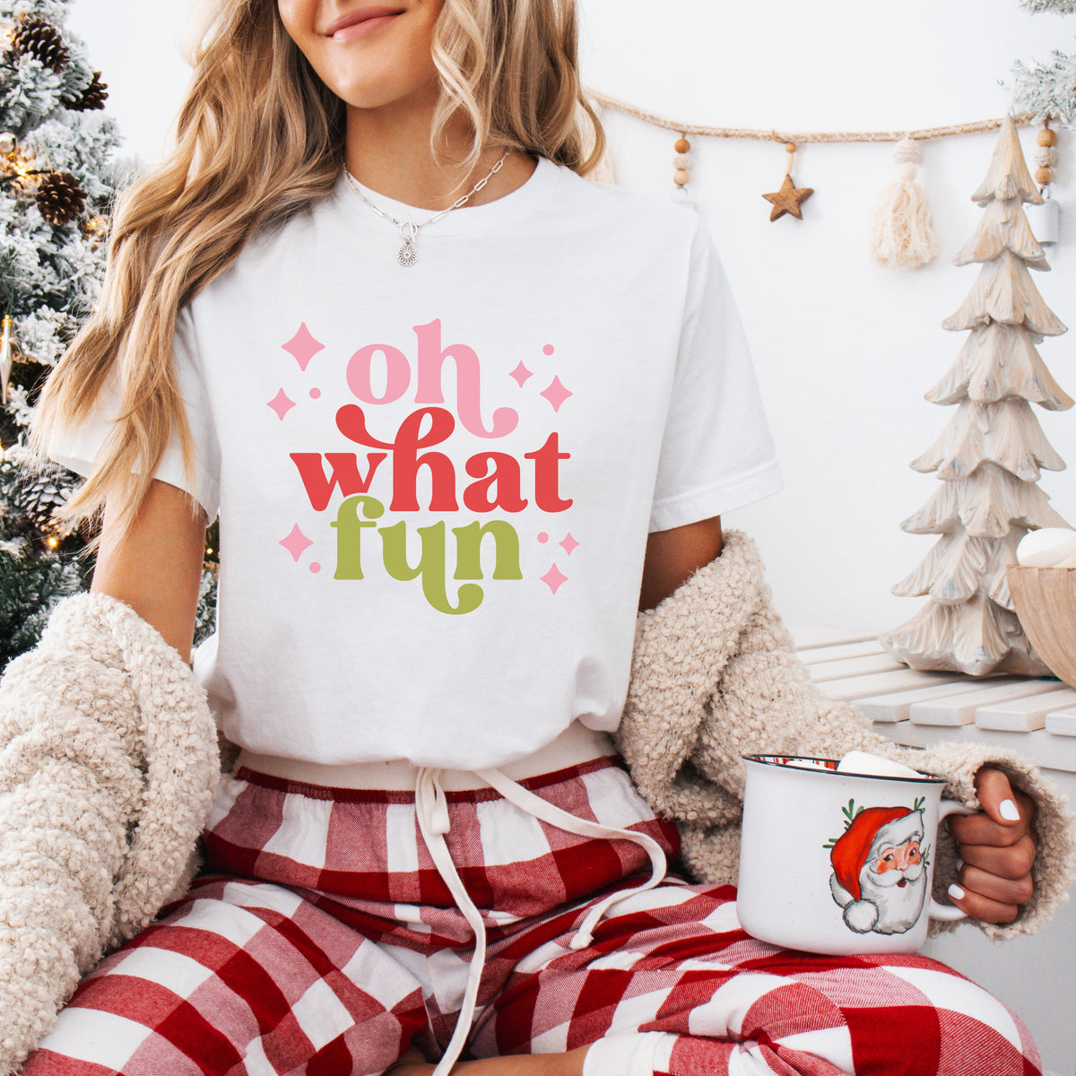 A person wearing a white t-shirt with the text &#39;Oh what FUN&#39; printed in pink and green colors, sitting in a cozy room with a Christmas tree and gifts in the background.