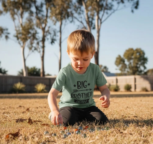 Young boy playing outdoors wearing a sage green Big Brother Again t-shirt with hand-lettered design and arrow detail