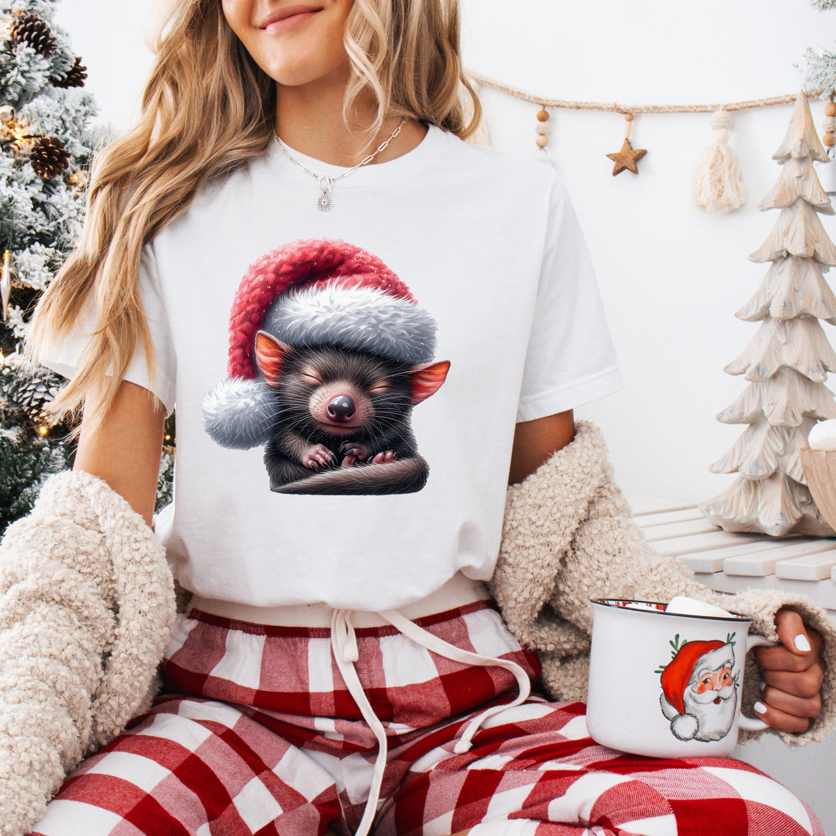 A person wearing a white Christmas-themed t-shirt with an illustration of a Tasmanian Devil wearing a Santa hat, sitting in a cozy room with a Christmas tree and gifts in the background.
