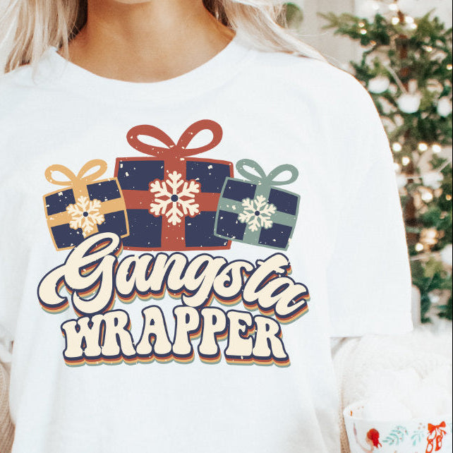 A person wearing a white sweatshirt with the text 'GANGSTA WRAPPER' printed on the front, holding a mug with 'BABY, KIDS &amp; ADULTS' text on it, in a cozy room with Christmas decorations.
