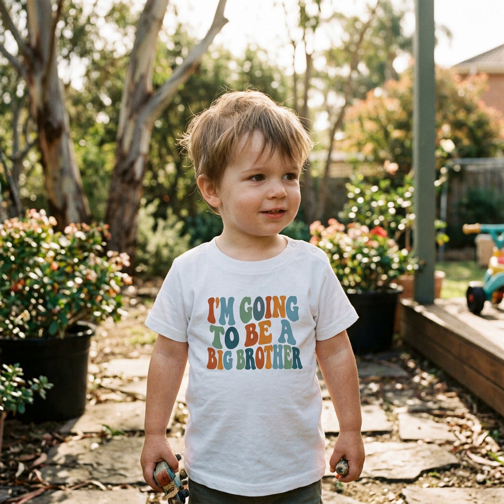 A white short-sleeve baby t-shirt with the phrase 'I'M GOING TO BE A BIG BROTHER' printed on the chest in a retro, wavy multicolor font.