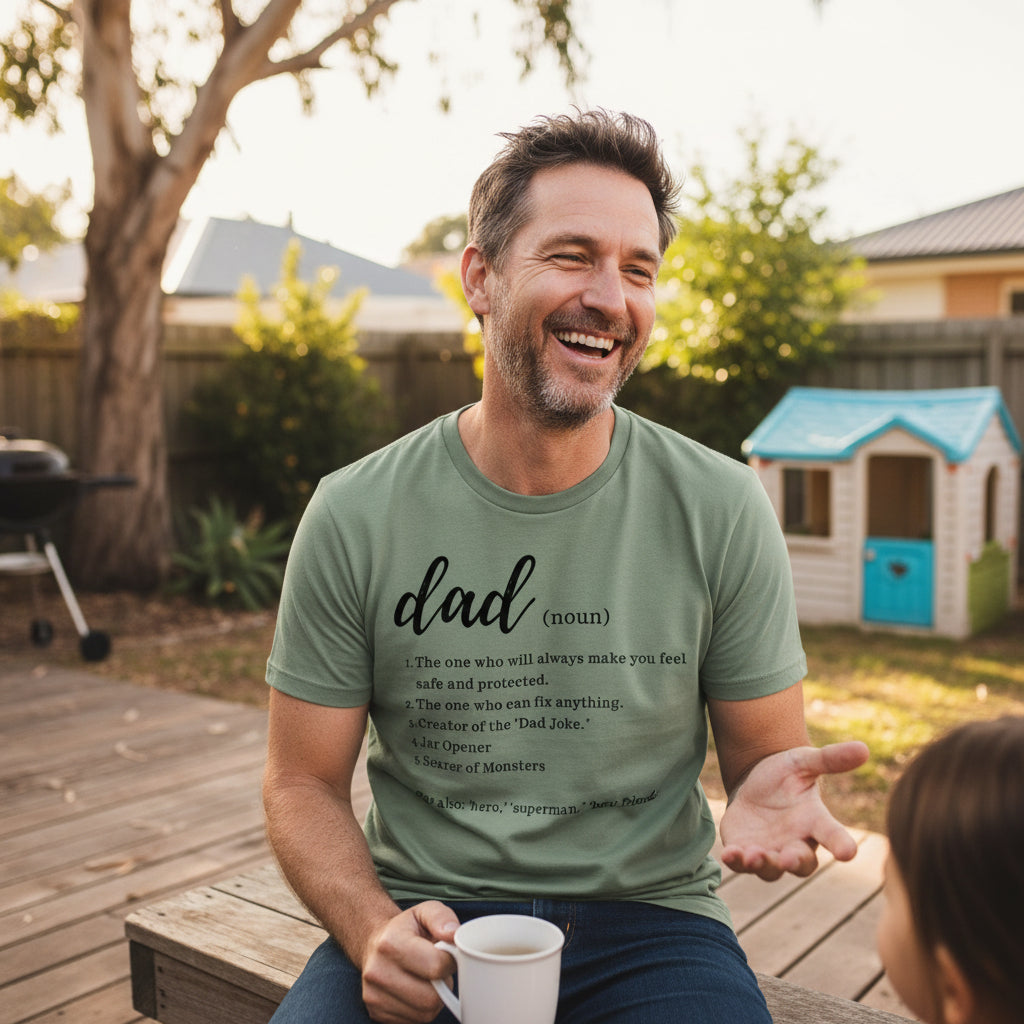 A man wearing a sage green graphic t-shirt with a humorous dictionary definition of 'dad' sitting on a wooden deck in a backyard, holding a white mug.